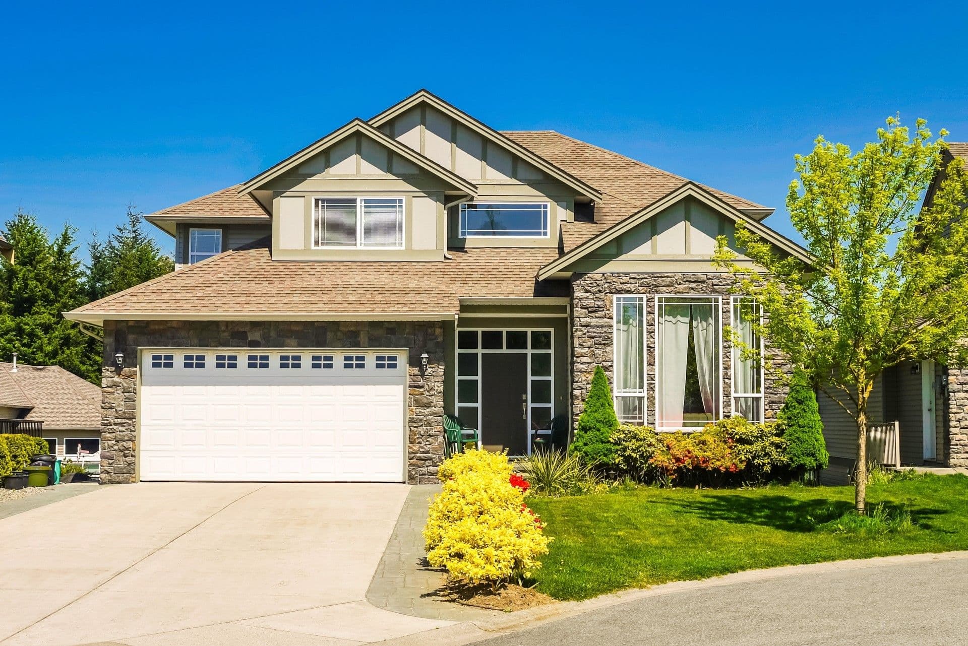 Concrete driveway - Two-story residential house with stone and beige siding, attached garage, multiple gables, and manicured landscaping including yellow shrubs and green lawn under clear blue sky
