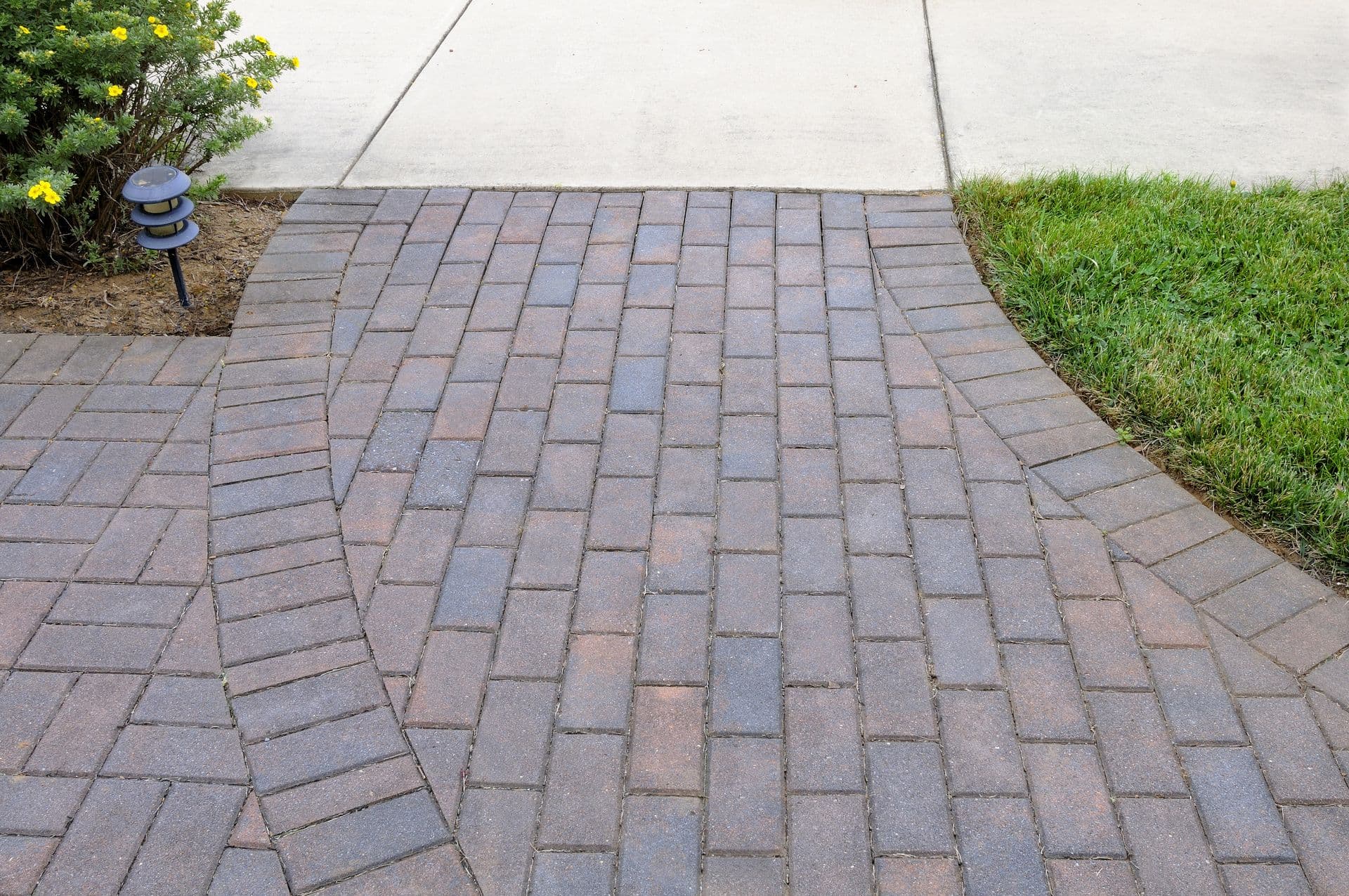 Brick paver driveway with herringbone pattern leading to concrete walkway, flanked by lawn and flowering shrub with solar light