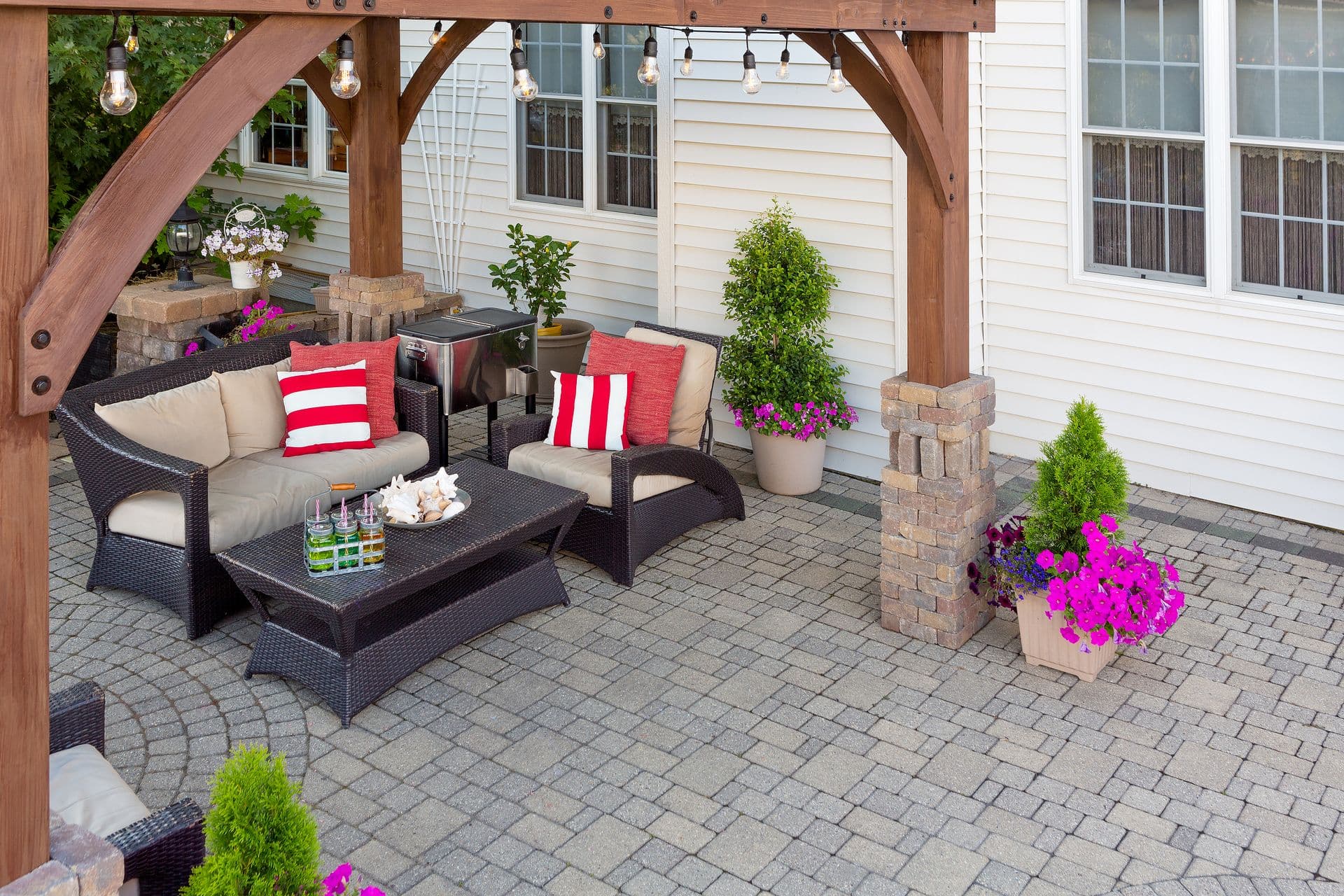 Outdoor patio with wooden pergola featuring string lights, black wicker furniture with beige cushions and red striped pillows, potted flowers and shrubs, and brick columns attached to a white house with windows