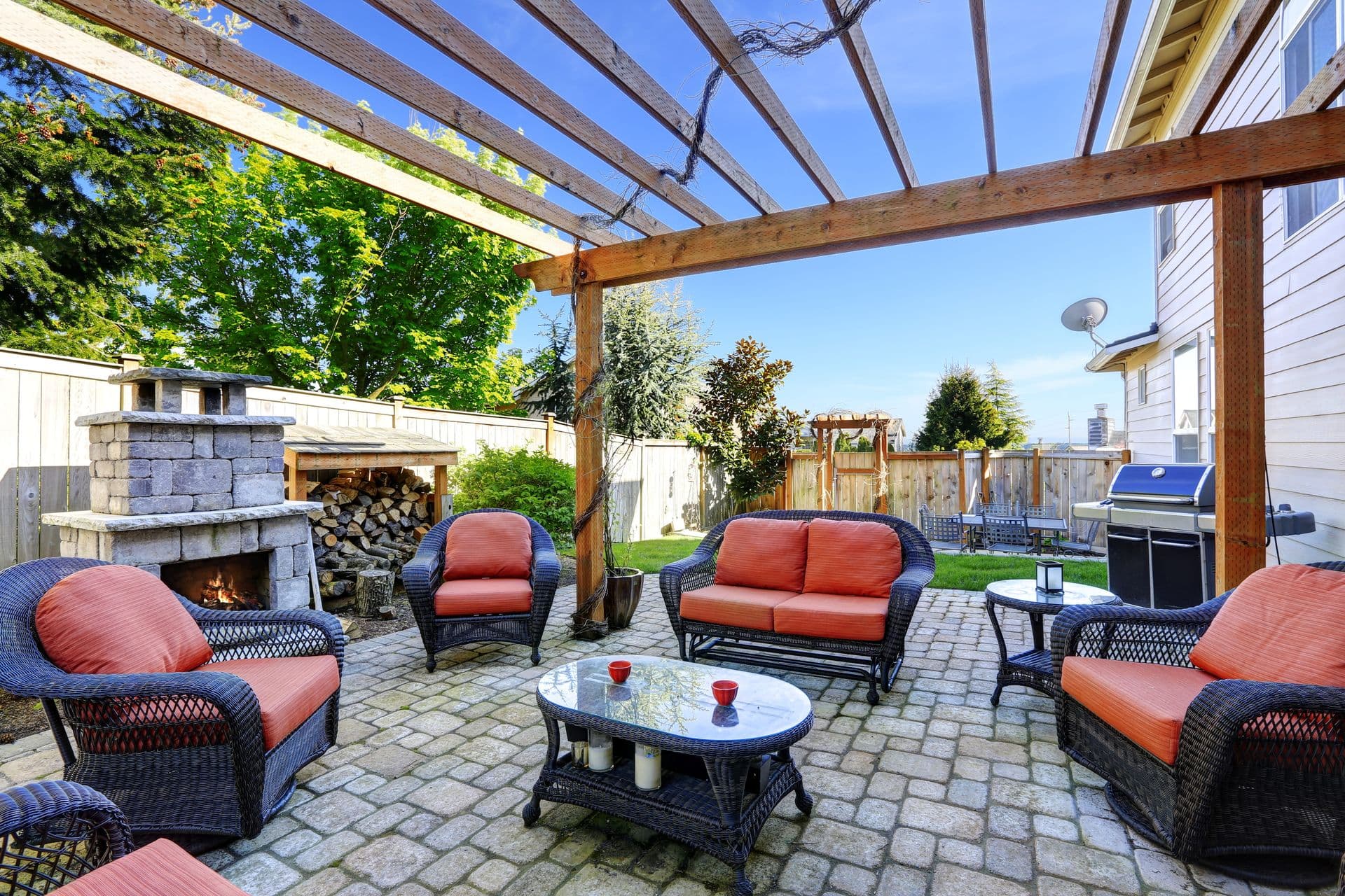 Spacious backyard patio with wooden pergola overhead, featuring wicker seating with coral cushions, stone fireplace, gas grill, and manicured lawn beyond