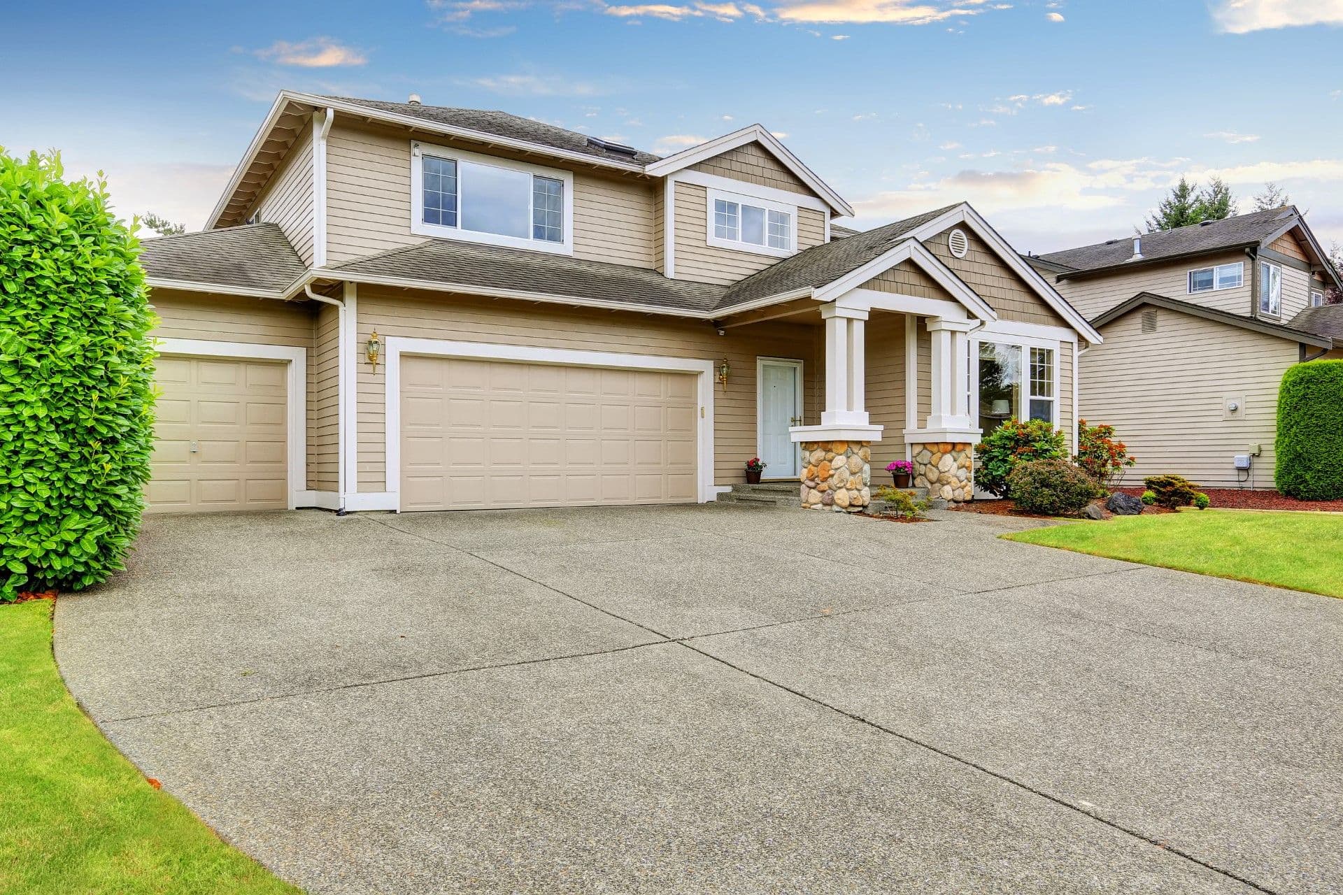Two-story beige house with tan and white trim, stone pillars framing the front entrance, double garage, and manicured lawn with green hedges, and concrete driveway