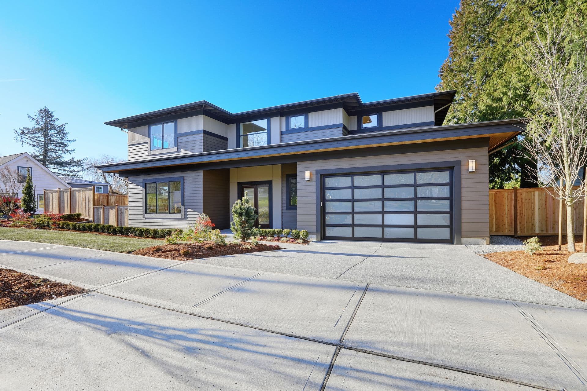 Modern two-story residential home with gray and white siding, large windows, dark garage doors, and landscaped front yard with concrete driveway