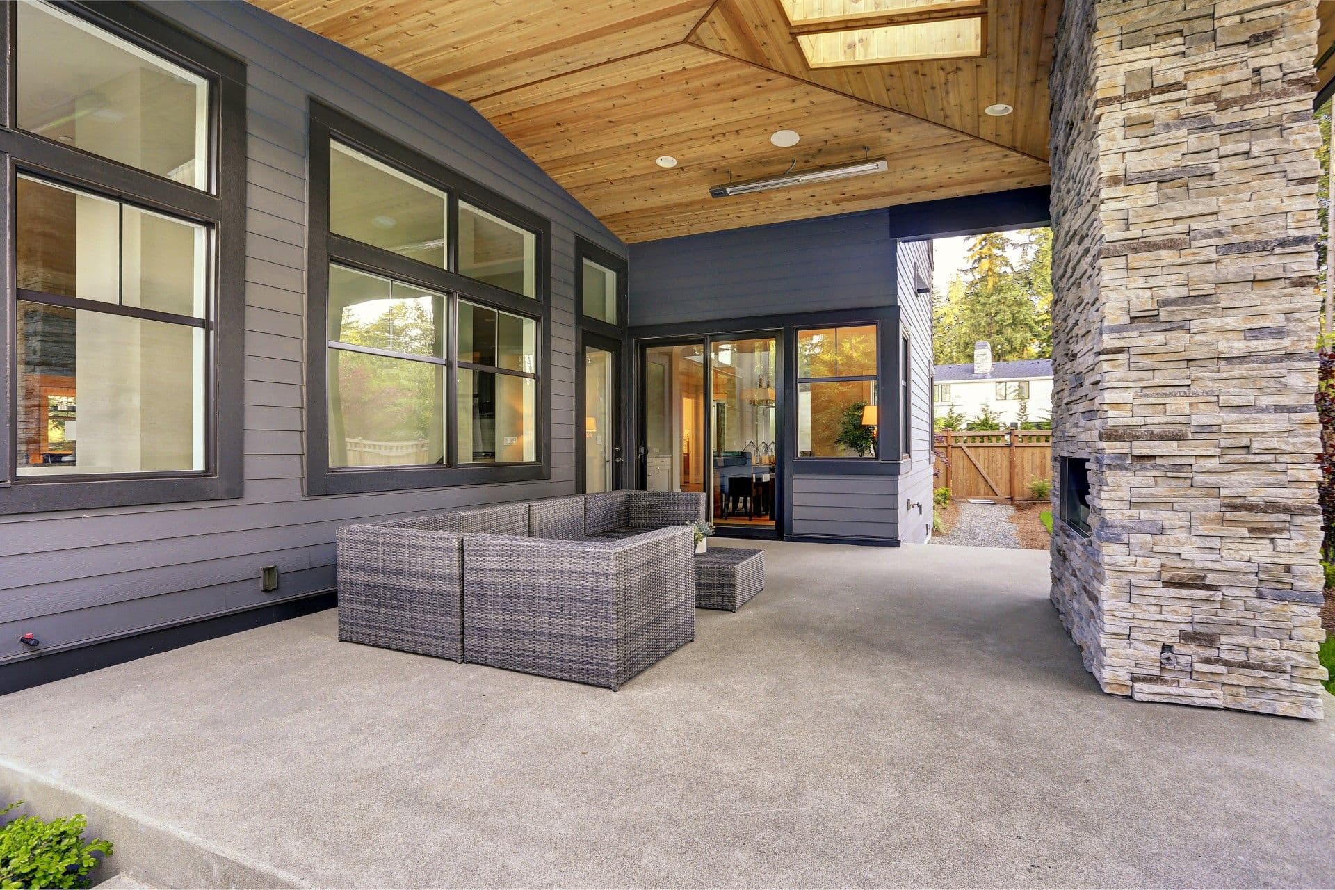 Modern covered patio with woven outdoor seating, black-framed glass doors and windows, wooden ceiling, stone pillar, and concrete flooring overlooking a backyard