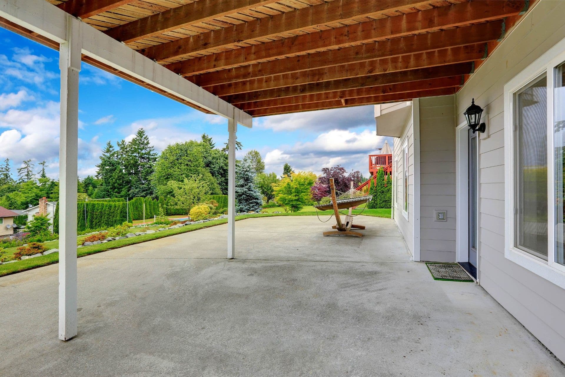 Covered patio with wooden beam ceiling, concrete floor, and white support columns overlooking a manicured backyard with hammock, landscaped gardens, hedges, and distant treeline