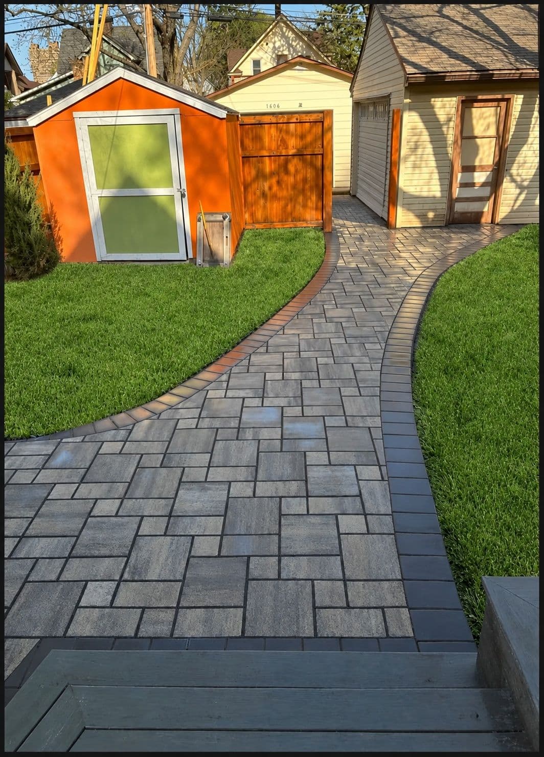 Backyard patio with herringbone paver pathway leading past an orange storage shed and cream-colored structures, flanked by green lawn and residential homes