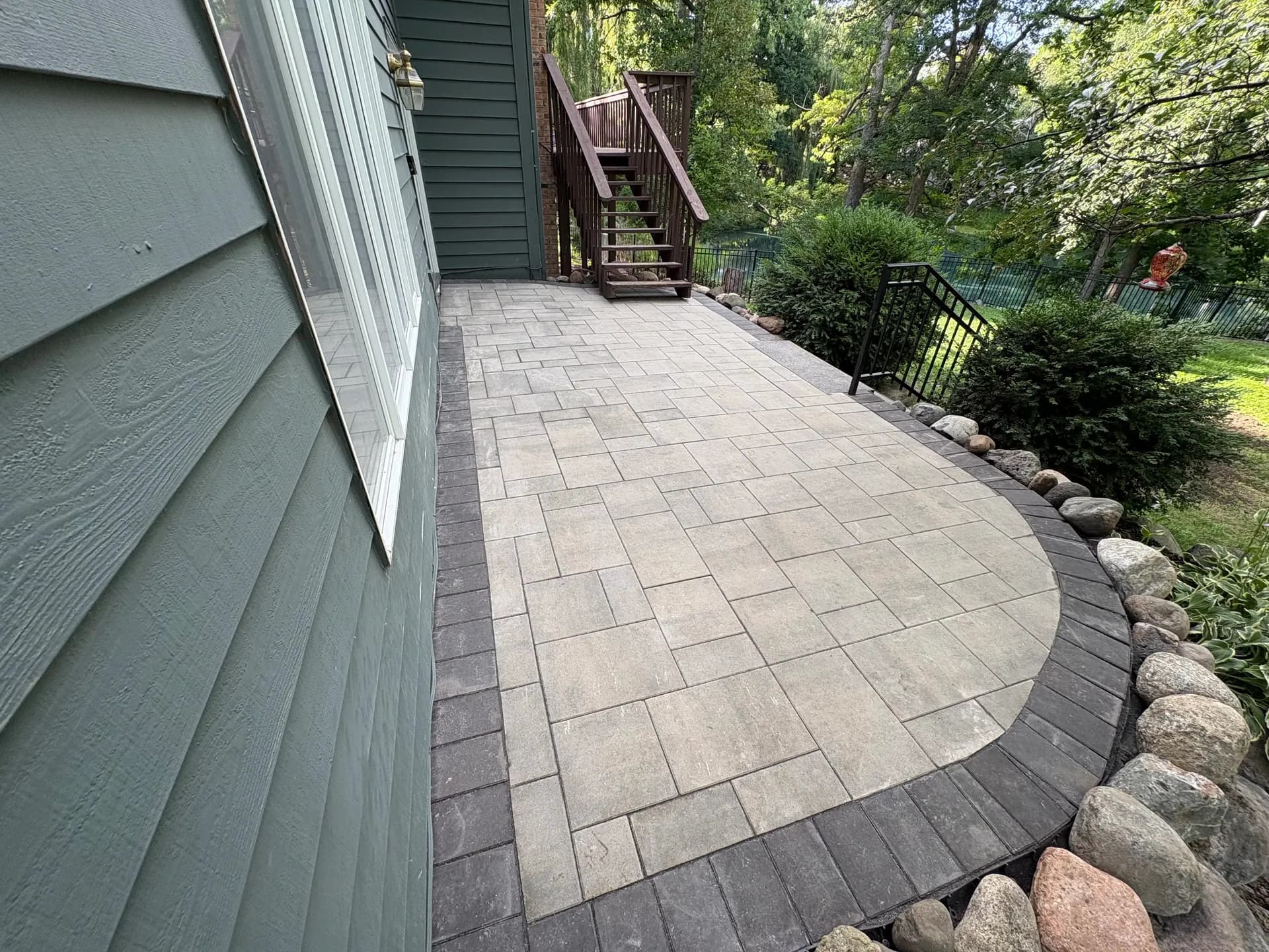 Paved patio with curved edge along house siding, wooden stairs and deck in background, surrounded by mature trees and landscaping with stone border edging