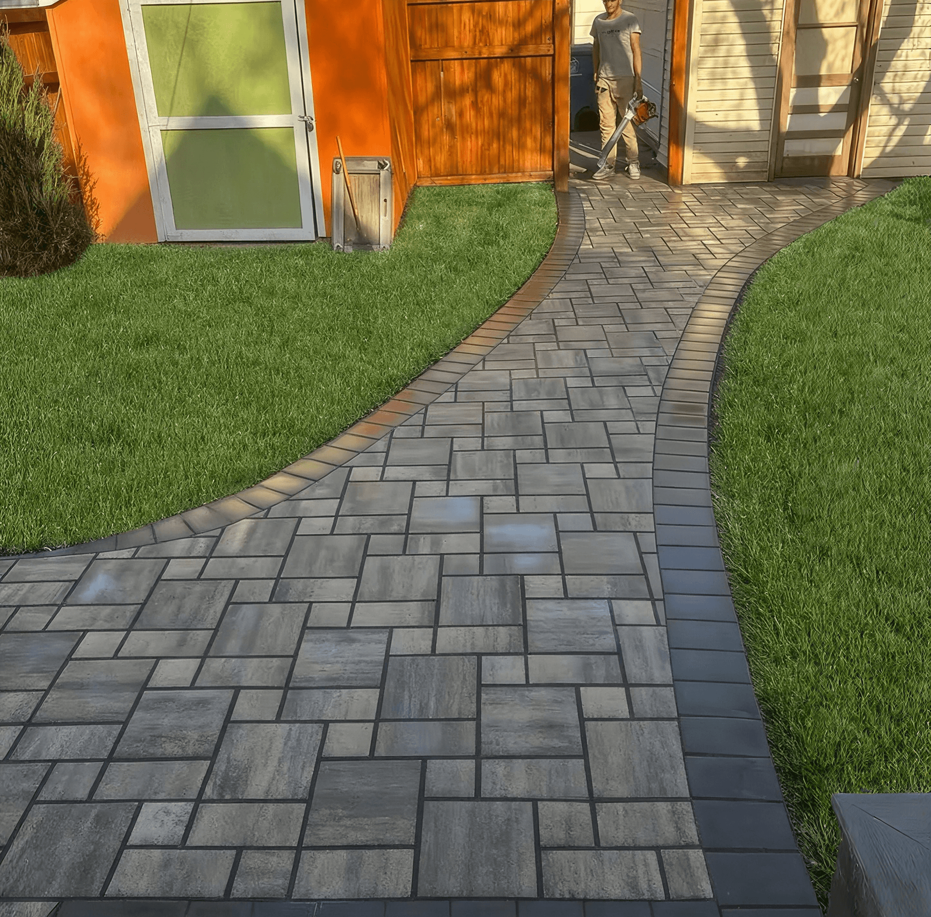 Curved paver pathway with herringbone pattern leading through modern residential yard with manicured lawn, orange modern building with glass door, and person standing in background